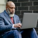 Mature African American businessman using a laptop while sitting outdoors, dressed in a suit.